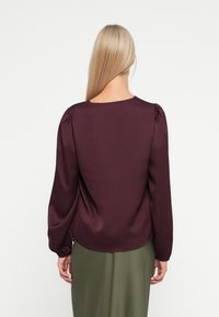 Bluză burgundy cu mâneci lungi și decolteu rotund. Material neted cu umăr bufant și tiv curbat. Asortată cu o fustă verde.
