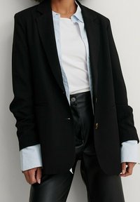 Blazer nero dal design sartoriale, con bottoni dorati, abbinato a una camicia button-up azzurra chiara e pantaloni in pelle nera.