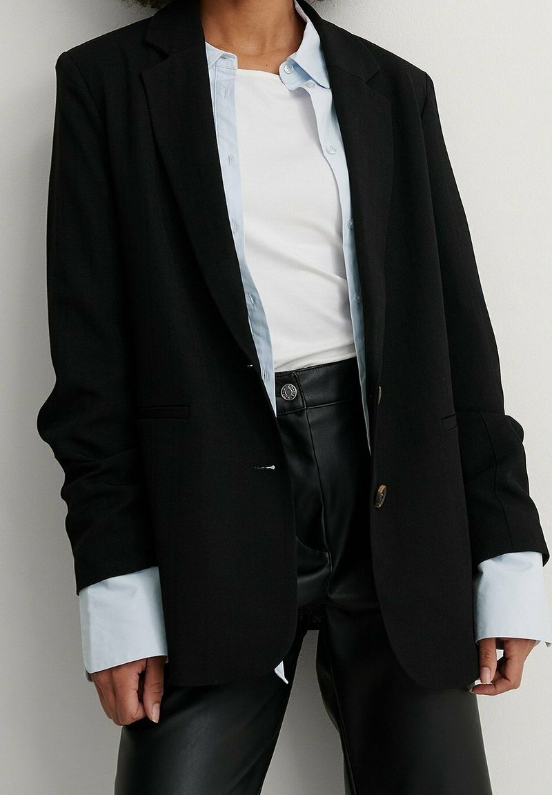 Blazer nero dal design sartoriale, con bottoni dorati, abbinato a una camicia button-up azzurra chiara e pantaloni in pelle nera.