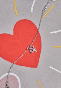 Pulsera de cadena de plata con un charm de cruz, un charm de corazón pequeño y una cuenta brillante. El fondo es un gran corazón rojo sobre gris.
