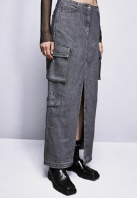 Gonna maxi in denim grigio con spacco frontale, dotata di due grandi tasche laterali e una tradizionale chiusura con zip. Abbinata a mocassini neri e calzini.