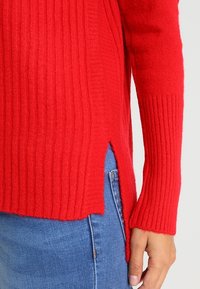 Punainen ribbiknit-sweateri pitkillä hihoilla ja sivuaukkoilla, valmistettu pehmeästä materiaalista. Yhdistetty vaaleansiniisiin farkkuihin.