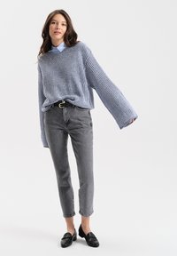 Blauwe gebreide trui met brede mouwen, gecombineerd met grijze high-waisted jeans en zwarte loafers, met daaronder een overhemd met klassieke kraag.