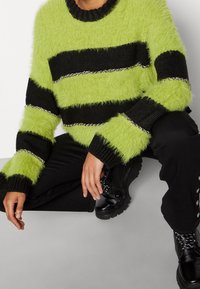 Vezeliger groene en zwarte gestreepte trui met zilveren kettingaccenten; ronde hals en brede mouwen. Model draagt zwarte broek en laarzen.
