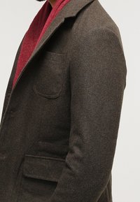 Brauner Mantel aus Wollmischung mit einem Reverskragen, Seitentasche und Knopfverschluss. Getragen über einer strukturierten roten Innenschicht.