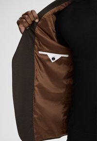 Brauner Blazer mit glattem Innenfutter. Verfügt über eine weiße Karteninnentasche mit Knopf. Die Außenseite besteht aus einem strukturierten, dunklen Stoff.