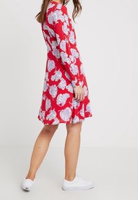 Vestido de tela roja con mangas largas, que presenta un patrón floral blanco. El vestido tiene un dobladillo acampanado y se combina con zapatillas blancas.