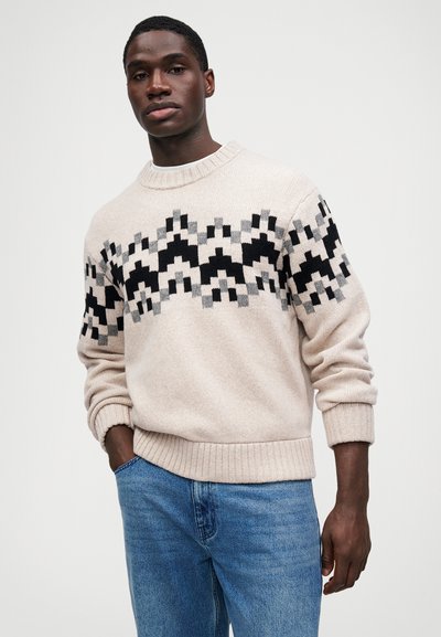Pulover tricotat bej cu modele geometrice negre și gri pe piept. Guler rotund, manșete și tiv cu margini ondulate. Asortat cu blugi denim albaștri.