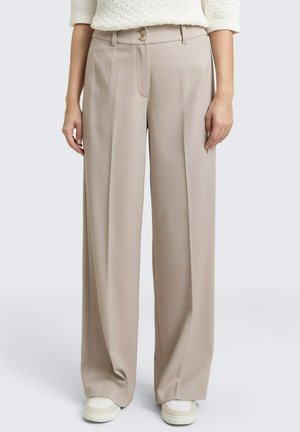Kobieta ubrana w beżowe spodnie o szerokich nogawkach z frontowymi plisami, szlufkami na pasek i podwójnymi drewnianymi guzikami, zestawione z białymi trampkami i kremowym swetrem dzianym.