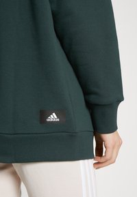 Sudadera verde oscura con puños y dobladillo acanalados, adornada con un parche del logo negro de Adidas en la parte inferior.