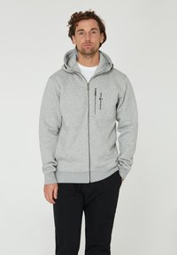 Sudadera gris con cremallera hecha de un tejido suave, con capucha, bolsillos laterales y un logo en el pecho. Acompañada de pantalones negros.
