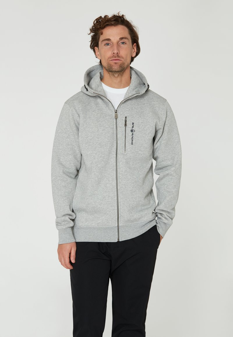 Sudadera gris con cremallera hecha de un tejido suave, con capucha, bolsillos laterales y un logo en el pecho. Acompañada de pantalones negros.