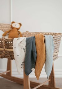 Čtyři bavlněné zavinovačky v bílé barvě s potiskem mořského koníka, tyrkysové, hořčicové a světle modré, přehozené přes tkaný koš s dřevěnými nohami.