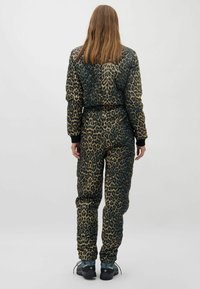 Person, der mit dem Rücken zur Kamera steht, trägt eine leopardgemusterte gesteppte Jacke und Hose sowie schwarze Wanderschuhe vor einem weißen Hintergrund.