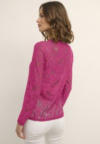 Top in pizzo fucsia con maniche lunghe, motivo floreale e texture trasparente. Presenta un colletto alto e un orlo scallopato, abbinato a pantaloni bianchi.