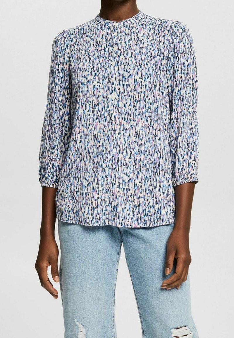 Blouse à motif floral avec manches trois-quarts, col rond, dans des tons de bleu, rose et blanc ; tissu doux et fluide. Portée avec un jean bleu clair.