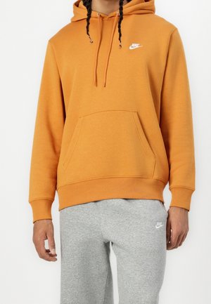 Homme portant un sweat à capuche orange Nike avec des cordons et une poche avant, associé à un pantalon de survêtement Nike gris clair.