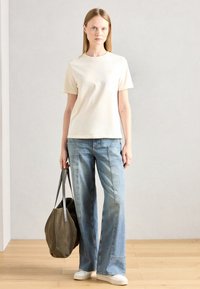 T-shirt in cotone beige con maniche corte, abbinato a jeans a gamba larga blu chiaro. Tiene una borsa tote in suede verde scuro. Indossa sneakers bianche.