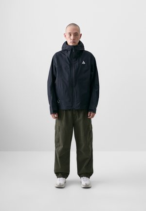 Veste noire imperméable avec capuche, fermeture éclair à l'avant, poches latérales et logo blanc. Associée à un pantalon cargo vert olive ample et des chaussures blanches.