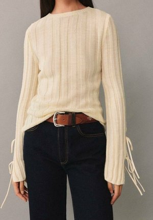 Mujer que lleva una camiseta larga de canalé color crema con cordones laterales, vaqueros oscuros de talle alto y cinturón de cuero marrón con hebilla plateada.