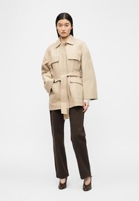 Γυναίκα που στέκεται φορώντας μπεζ σακάκι με ζώνη και μεγάλα τσέπες, σκούρο καφέ παντελόνι και καφέ γοβάκια, σε απλό λευκό φόντο.