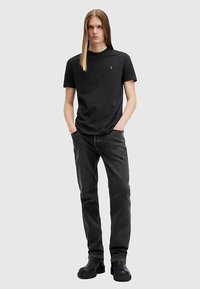 Melns apaļas apkakles T-krekls ar izšūtu logotipu, kombinēts ar tumšām džinsa biksēm. Modelis stāv ar rokām kabatās, valkājot melnas zābakus.