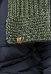 Écharpe en tricot verte avec un design texturé et côtelé, agrémentée d'une étiquette en cuir tissé. L'arrière-plan présente un tissu noir rembourré.