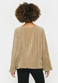 Haut à manches longues plissé dans une teinte beige avec un col rond et un ourlet festonné. Présente une texture verticale et une coupe décontractée.