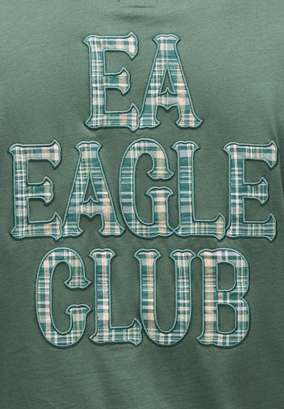 Πράσινο ύφασμα με μεγάλα ραμμένα γράμματα σε πράσινο και μπεζ καρό που σχηματίζουν τη φράση «EA EAGLE CLUB» οργανωμένα σε τρεις γραμμές.