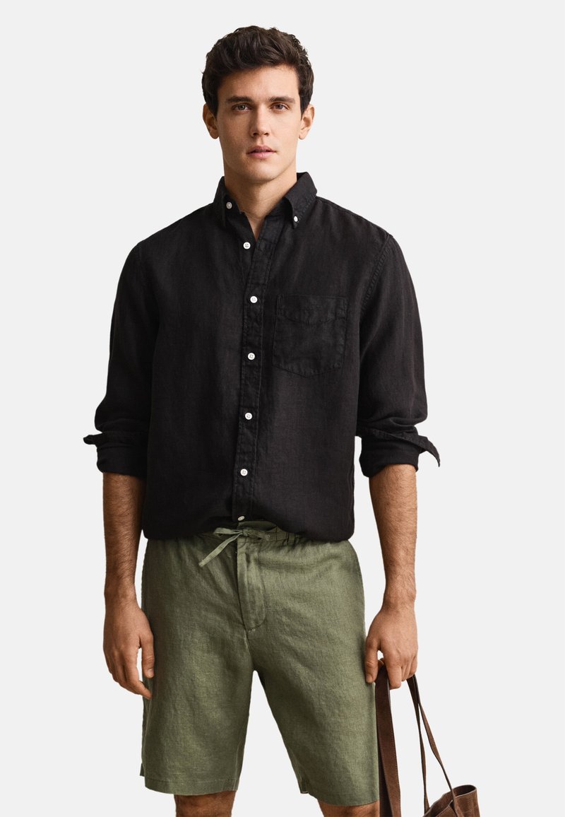 Homme portant une chemise noire boutonnée avec manches retroussées et un short vert à cordon, tenant un sac fourre-tout marron à côté.