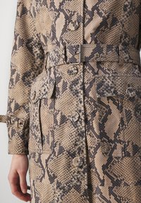 Manteau à motif peau de serpent beige et brun foncé, avec de gros boutons, un détail ceinture, et des poches avant avec rabats.