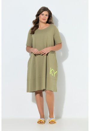 Kvinde iført olivengrøn knælængde kjole med gul "KY" tekst og blå og gule sandaler mod hvid baggrund.