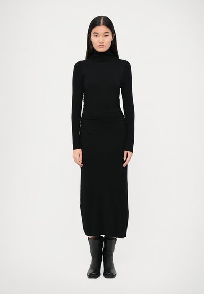 Fekete garbó pulóver és sima, rugalmas anyagból készült testhezálló maxi szoknya, fekete térdig érő csizmával párosítva. Minimalista dizájn.
