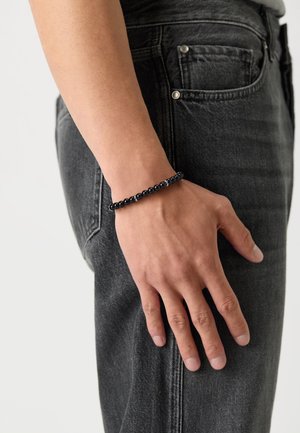 Schwarzes Perlenarmband am Handgelenk, mit glatten runden Perlen. Tragen auf einer Hand, die auf dunkelgrauen Jeans ruht. Einfaches und minimalistisches Design.