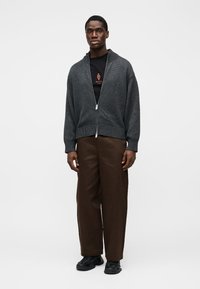 Sveter na zips v tmavosivej pletenine, voľný strih. Kombinovaný s hnedými nohavicami s širokými nohavicami a čiernymi teniskami, nosený na čiernom tričku.