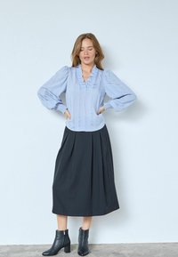 Šviesiai mėlyni marškiniai su raukšlėtu iškirpte ir pūstomis rankovėmis, derinami su juoda midi sijonu ir juodais kulkšnių batais su tekstūruotu paviršiumi.