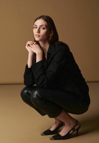 Sort langærmet skjorte, sorte læder cropped bukser og sorte højhælede sko med guld tåhætte. Modellen squatter foran en brun baggrund.