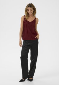 Bordo top s čipkastim detaljima na izrezu, u kombinaciji s crnim širokim hlačama i crnim cipelama s oštrim vrhom. Glatka tekstura tkanine.