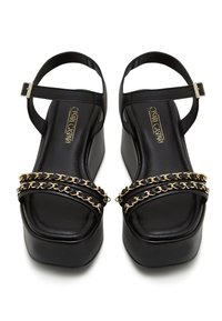 Sandali platform neri con una texture in pelle liscia, caratterizzati da due dettagli in catena dorata sulla parte anteriore del piede. Cinturino alla caviglia regolabile con fibbia.