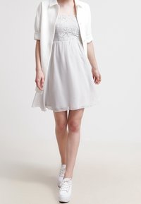 Robe blanche avec haut en dentelle et jupe évasée, superposée à une chemise blanche. Le mannequin porte des baskets blanches. Tissu lisse ; sans motifs apparents.