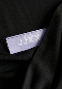 Svart tyg med en slät struktur, med en lavendelfärgad etikett sydd i, som visar bokstäverna "JJXX" i vit broderi.