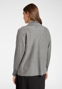 Pulóver gris jaspeado con cuello alto y mangas largas. Presenta una cinturilla ajustada y un sutil detalle de logo en la parte posterior. Textura suave.