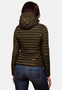 Doudoune vert olive avec une texture matelassée, capuche, et un patch de marque dans le bas du dos, associée à un jean bleu foncé.