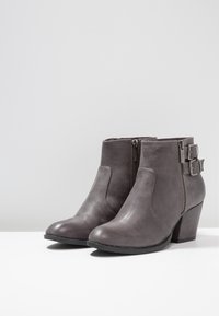 Botas de tobillo en imitación cuero gris con cremallera lateral y acentos de doble hebilla, presentando una punta redonda y un tacón apilado, mostradas sobre una superficie blanca.