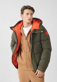 Chaqueta acolchada en verde oscuro con un forro de capucha en naranja brillante. Presenta un cierre de cremallera y botones a presión, combinada con capas interiores en tono beige.