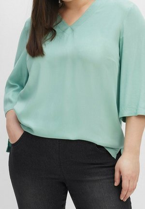 Helle Teal-Bluse mit V-Ausschnitt und drei Viertelärmeln, aus einem glatten Stoff gefertigt. Kombiniert mit dunklen Jeans aus Denim, die seitliche Taschen haben.