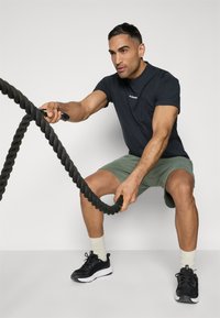 Black athletic t-shirt and green shorts worn by a man using a thick, twisted black battle rope, paired with black sneakers and beige socks.