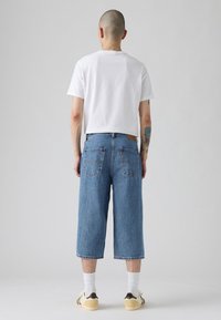 Culottes de mezclilla azul con un ajuste relajado, que cuentan con bolsillos traseros, un parche de cuero marrón y un dobladillo deshilachado, combinados con una camiseta blanca y zapatillas.