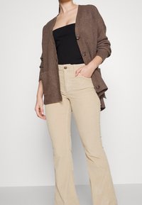 Brun ribbad cardigan med stora knappar, bärs över en svart topp och matchas med beige jagsbyxor med vida ben, vilket ger en avslappnad passform.