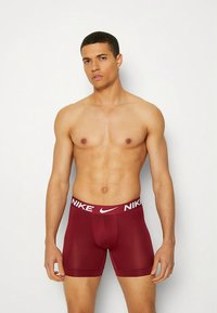 Moške bokser hlačke bordo barve z belim pasom z logotipom Nike, oprijetim dizajnom, gladko teksturo tkanine in dolžino do sredine stegna.
